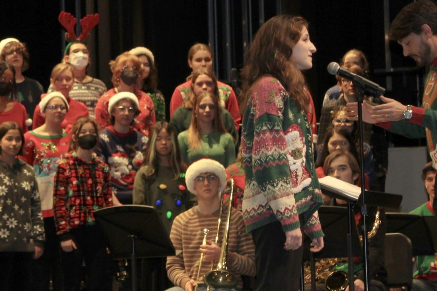 Female student wearing a christmas sweater stands behind microphone stand. Students in background wear a variety of holiday clothing and accessories.