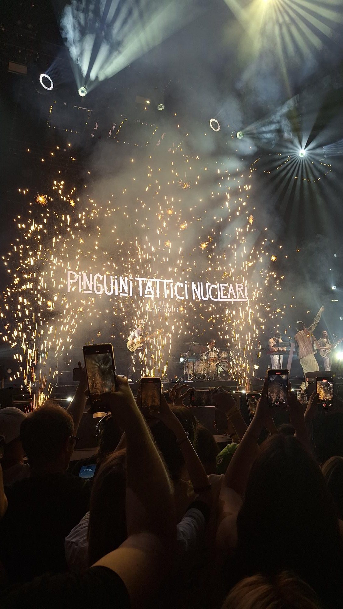 Crowd with hands up in the dark, gold sparks from the stage and a screen with Pinguini Tattici Nucleari written on it in white.