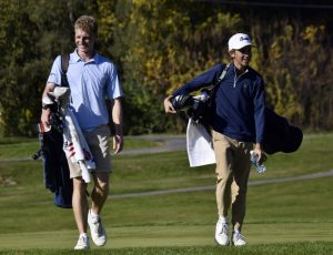 Sophomore Luke McGraw walks alongside a player from another team on the golf course. They both carry bags full of golf clubs. 