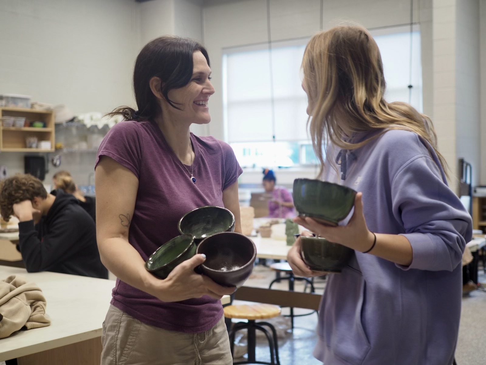 Two women are holding three bowls in their arms while laughing and looking at each other.