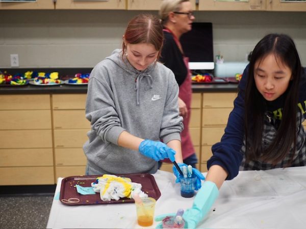 Seniors Mia Hollobaugh (left) and Emma Chen (right) during the art club tie-dye event on Tuesday,Nov. 19. They are standing at a desk, with a t-shirt on a maroon tray, and using liquid textile paint to color it.