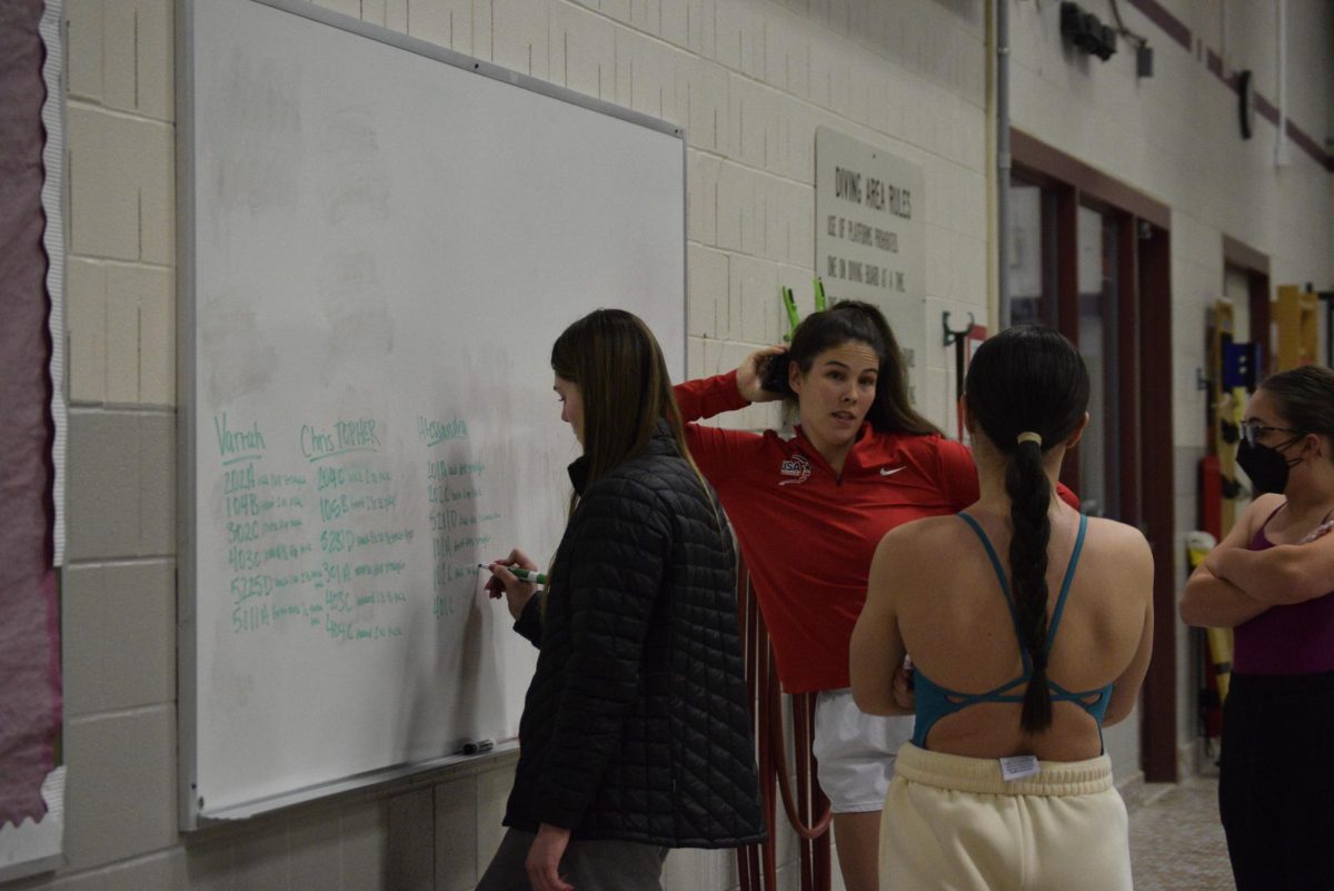 State High Junior Varrah Gramling writes on the whiteboard the dives that each team member will work on during practice, on Jan 8, 2025. In the background, coach Sierra Schomburg, freshman Cara Schuchert and junior Josephine Brannick discuss the upcoming practice. It’s Coach Schomburg's first season coaching, and she always brings a lot of energy. “Sierra, our coach is awesome. She's great. She's good at communicating, and she's great” Julia Johaenning, State High junior, and first year diver, stated.