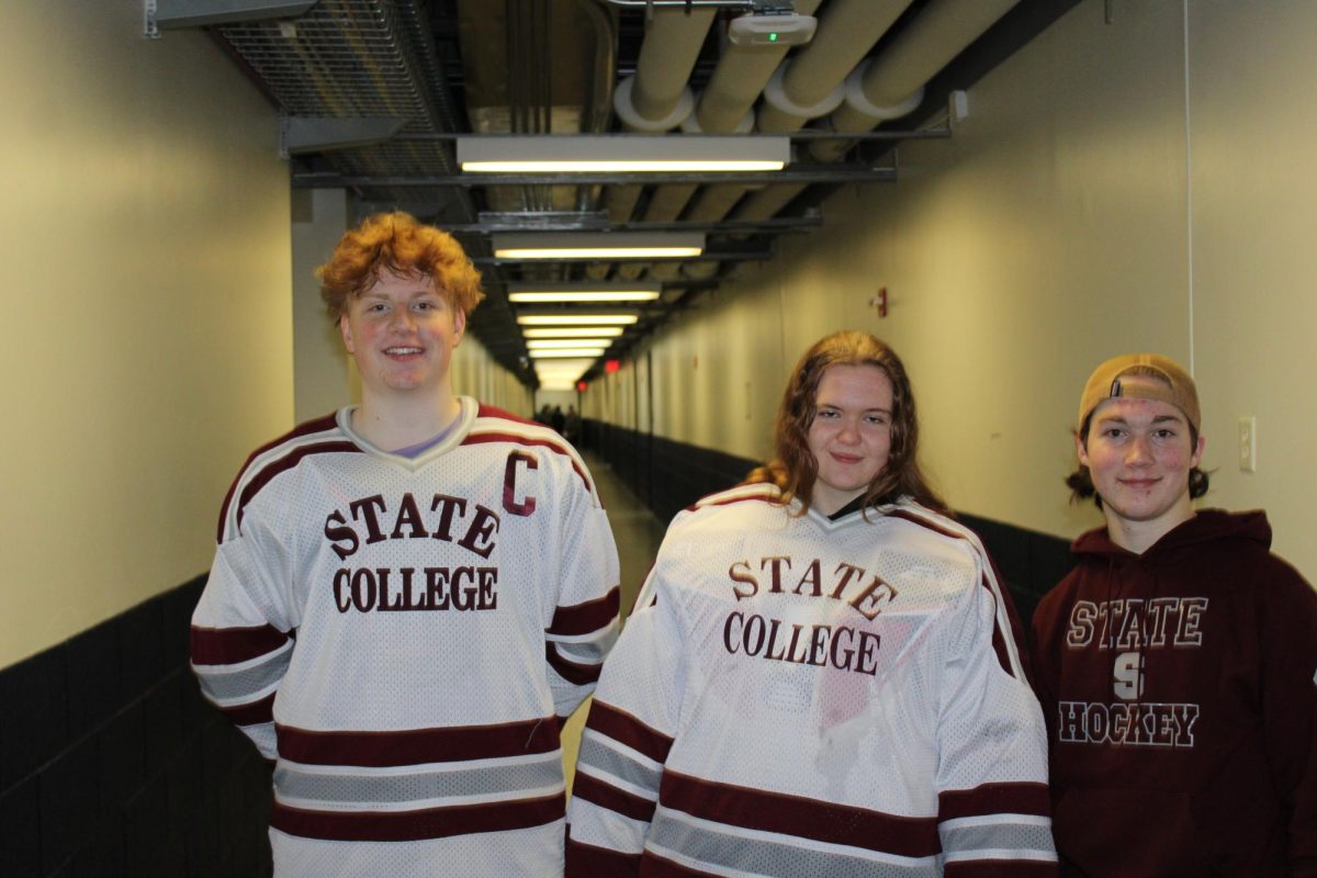 Seniors (left to right) Sean Murphy, Cecilia Keys, and Cooper Brumberg pose in an arena hallway after the game. Murphy and Keys wear their white home uniforms with State College on the front with maroon and grey striping while Brumberg wears a maroon State Hockey sweatshirt.