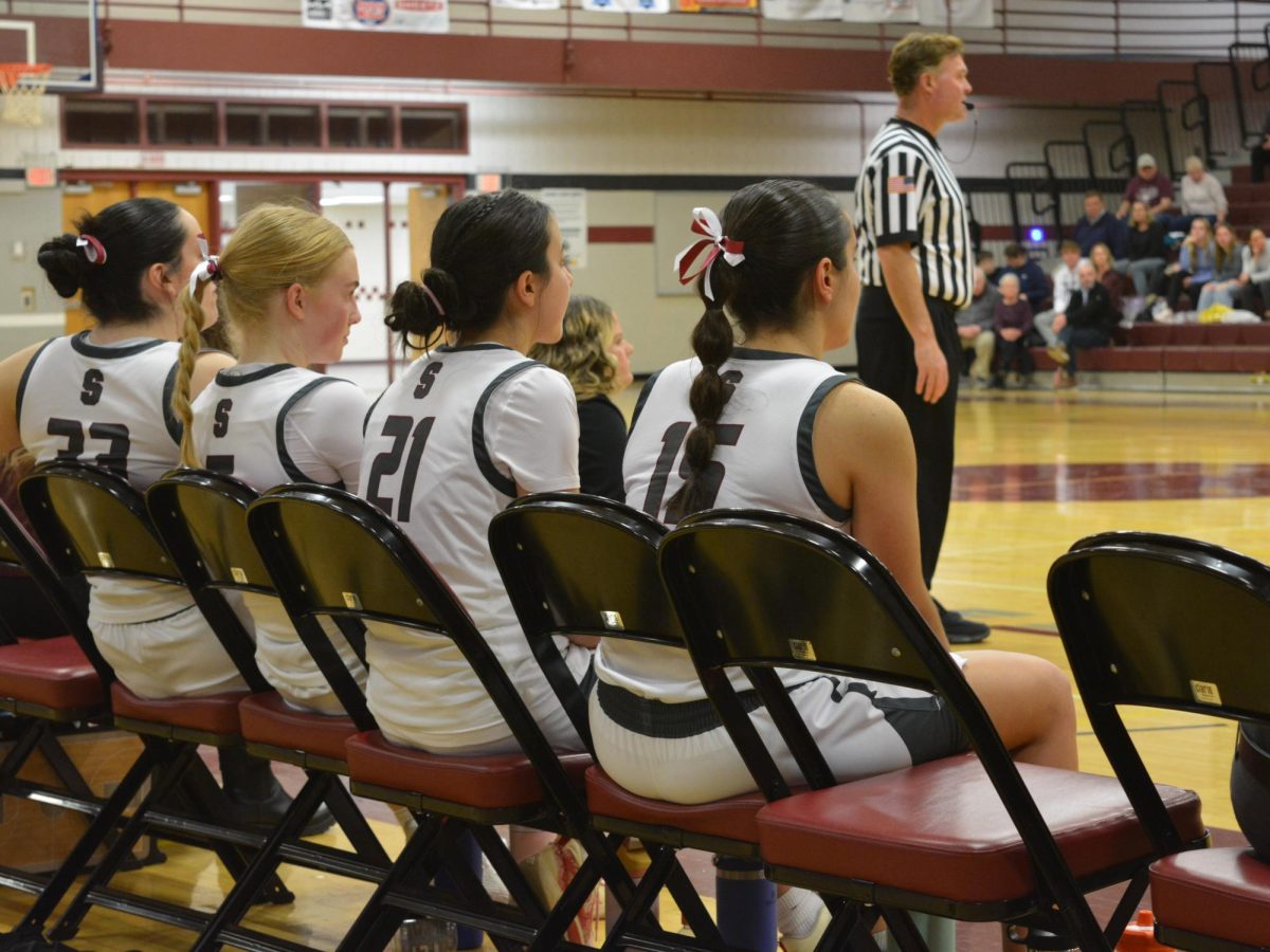 Seniors Risha, Bechtel, Irmak, and Roan sit together on the bench, cheering on their teammates and awaiting their call back to the court. They are all wearing maroon and white ribbons in their hair to commemorate Senior Night.
