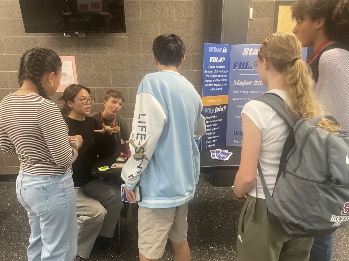 Students gather around a table at the club fair as club officers persuade them to join.