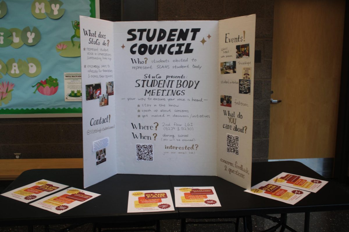 Student Council member sits by a poster for the club and tells students that they should join the club to help their voices be heard.