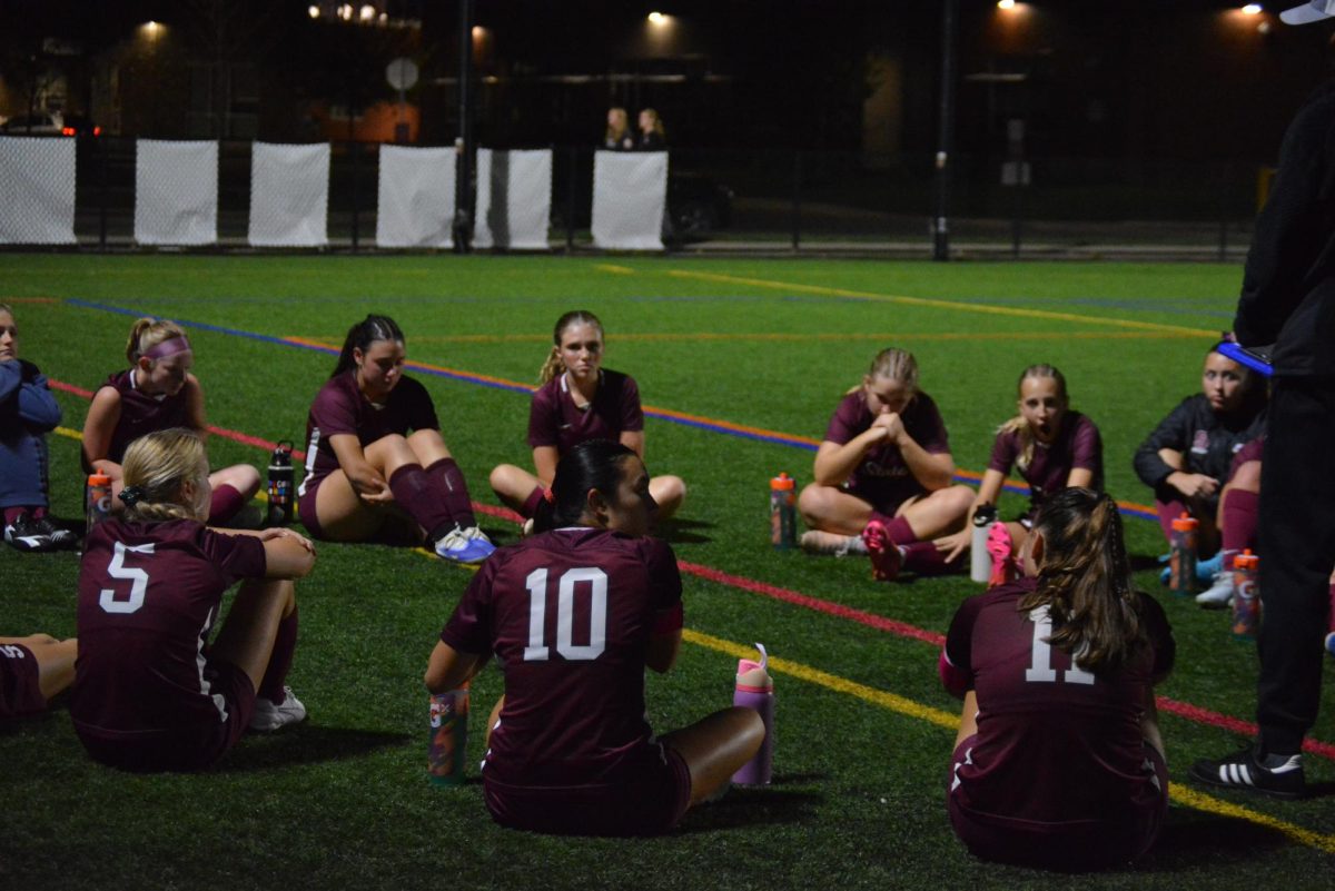 Senior Leila Crandall (#10) and some of her other teammates sit down in a circle during halftime as Head Coach Kent Dwyer talks about the game and what they need to focus on during the second half. Coach Kent Dwyer asks them their feedback as they reflect on the first half. 