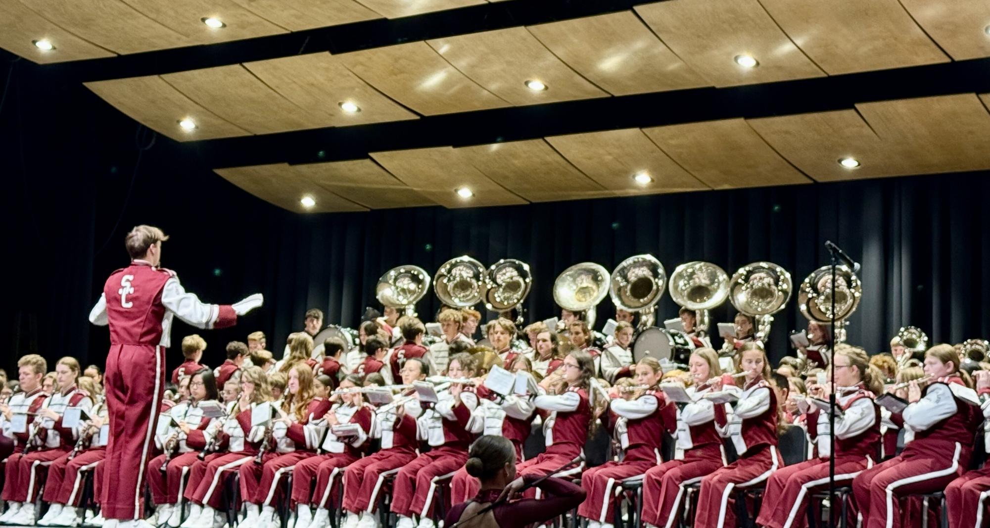 Assistant Drum Major Lewis Gipe stands facing the Marching Band, with his arms extended. He is located near the left of the screen. Many marching band members are sitting in chairs, facing forward with a variety of instruments.