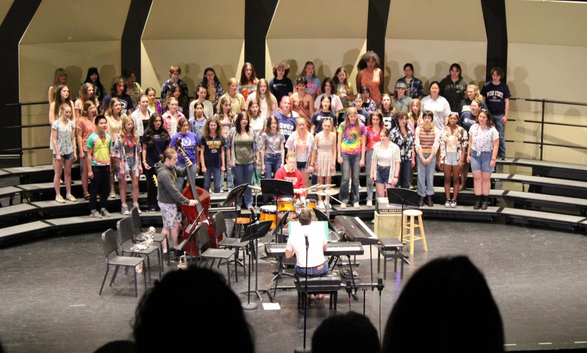 The choir performs “From the Start” in front of a crowd in the performing arts center. 
